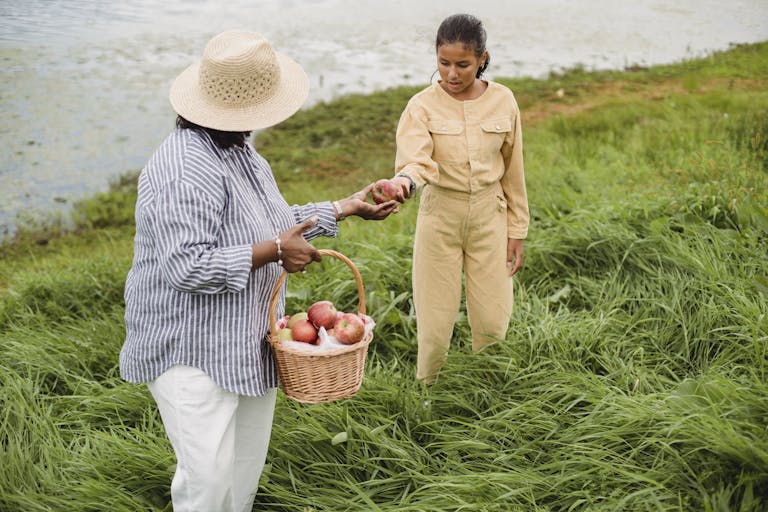 Unrecognizable ethnic woman carrying wicker basket with apple harvest and giving fresh fruit to girl standing in high grass near lake