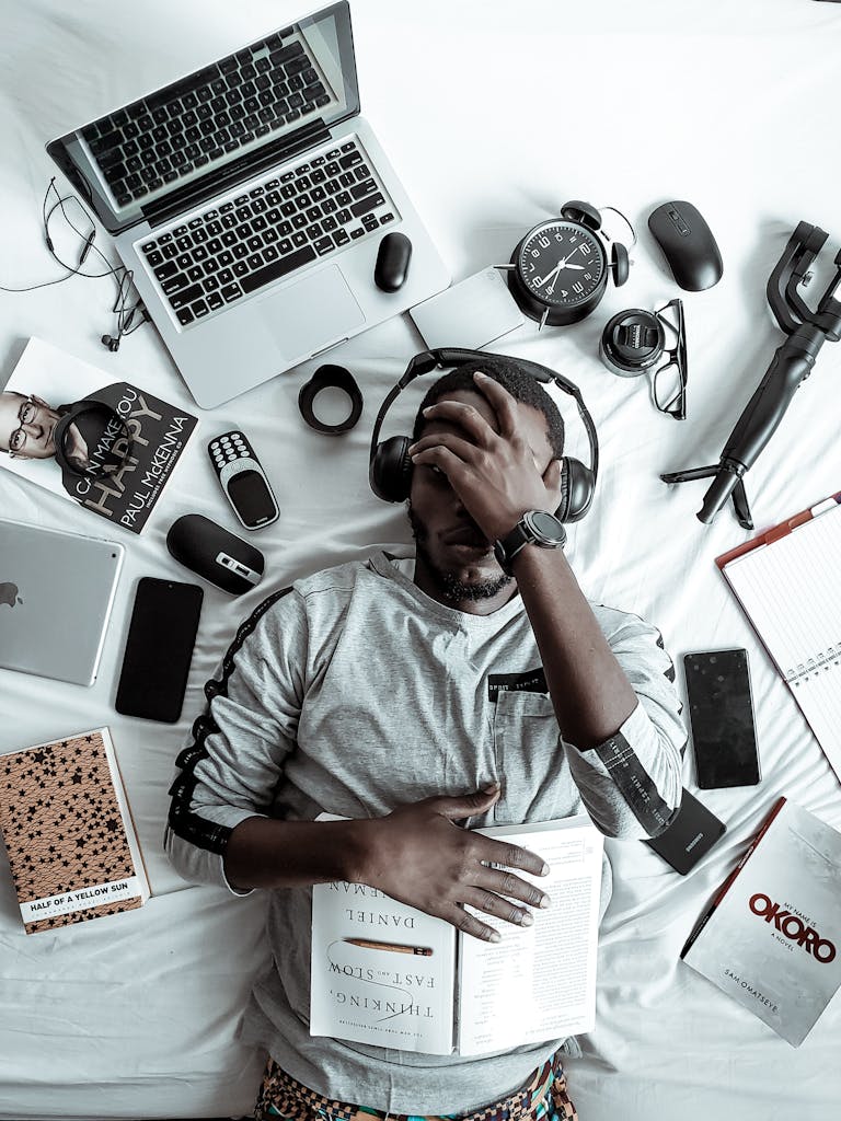 A young man lying on a bed surrounded by modern gadgets, books, and tech devices, illustrating digital lifestyle.